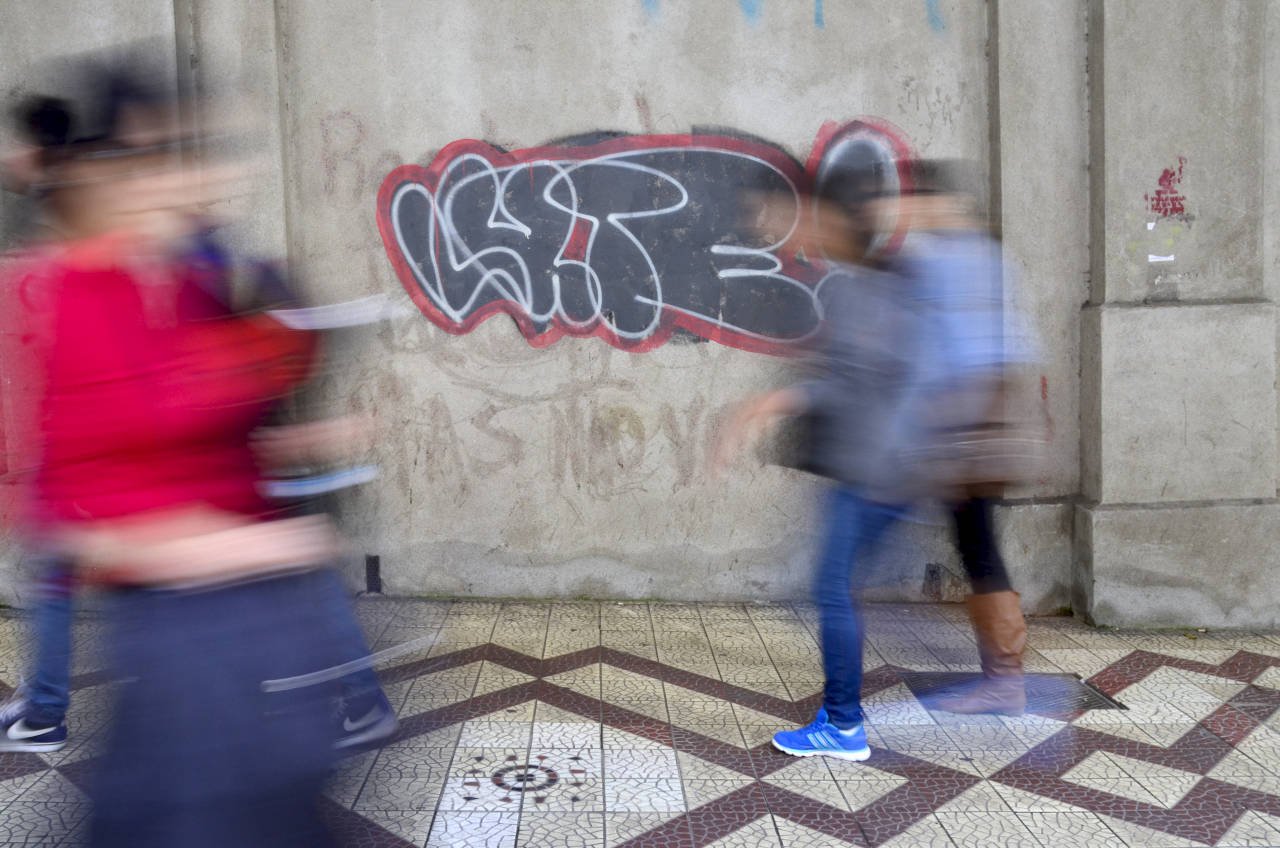 Pessoas em movimento borrado caminhando em frente a uma parede com grafite, mostrando uso de velocidade baixa na fotografia urbana para ilustração de técnicas de fotografia.
