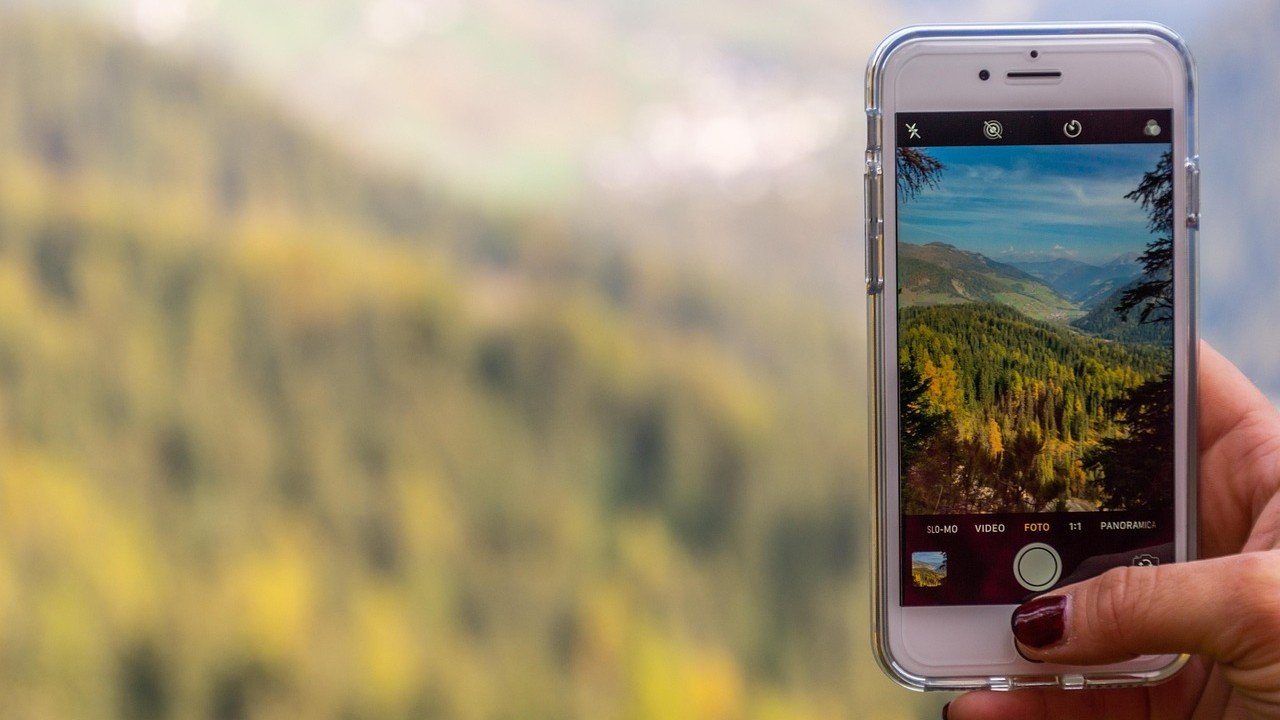 Mão segurando celular fotografando paisagem de montanha coberta por árvores, ilustrando artigo 3:4 ou 4:5 no Instagram?