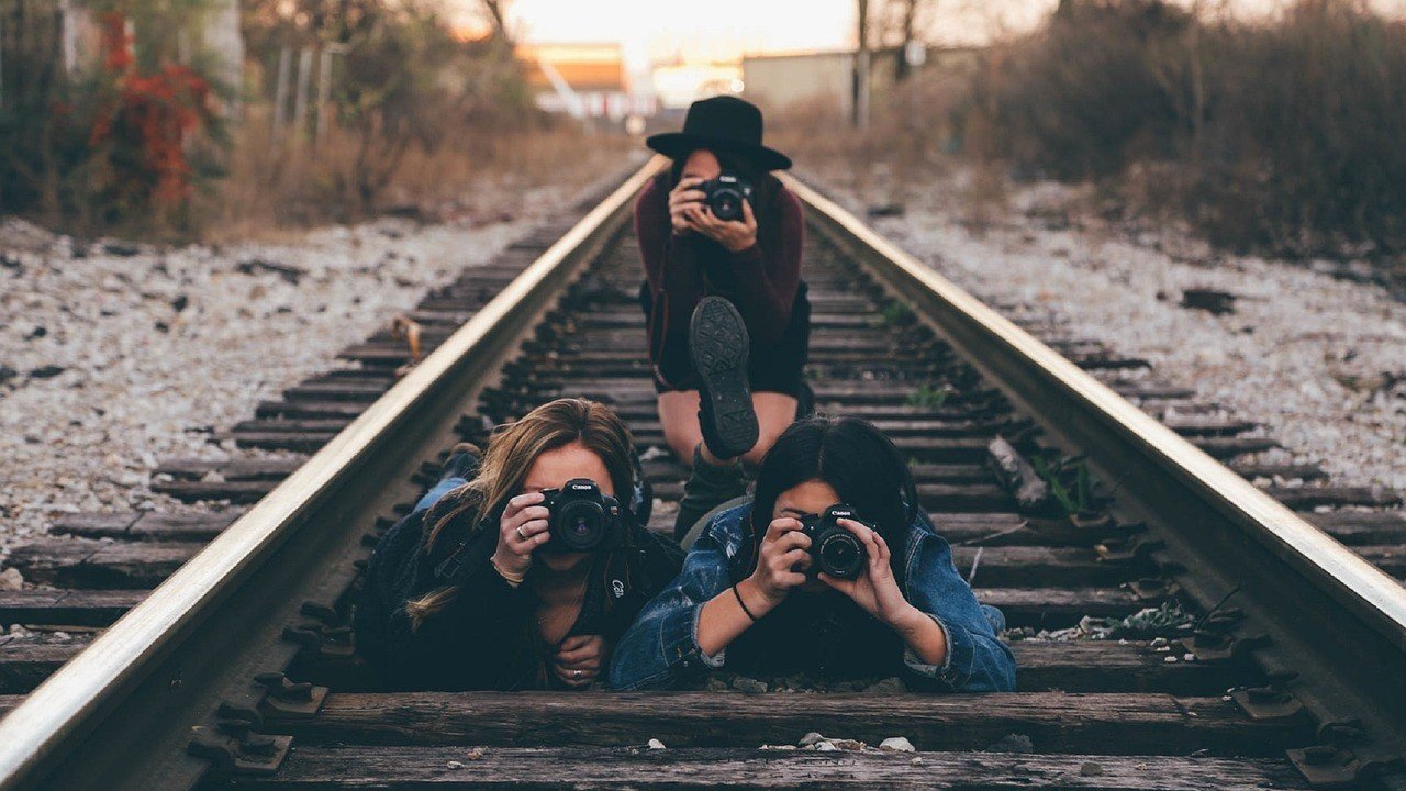 Três pessoas portando câmeras sobre um trilho de trem, ilustrando artigo Qual tipo de câmera faz sentido hoje: DSLR, Mirrorless ou compacta?