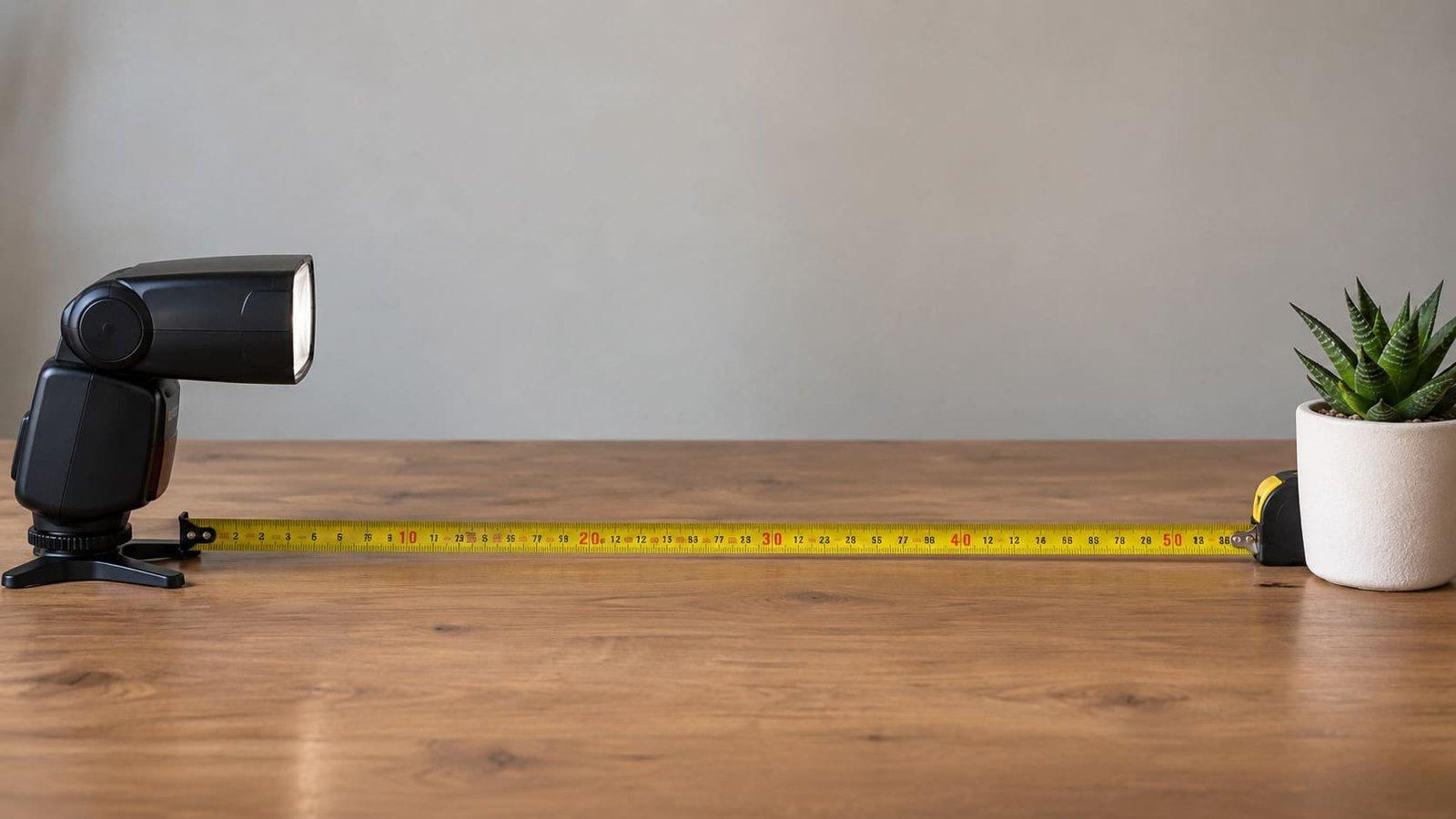 Flash èsquerda, sobre a mesa e um vaso com folhagem à direita, uma trena entre eles, ilustrando artigo Número guia do flash: como saber se ele alcança a distância de que você precisa.